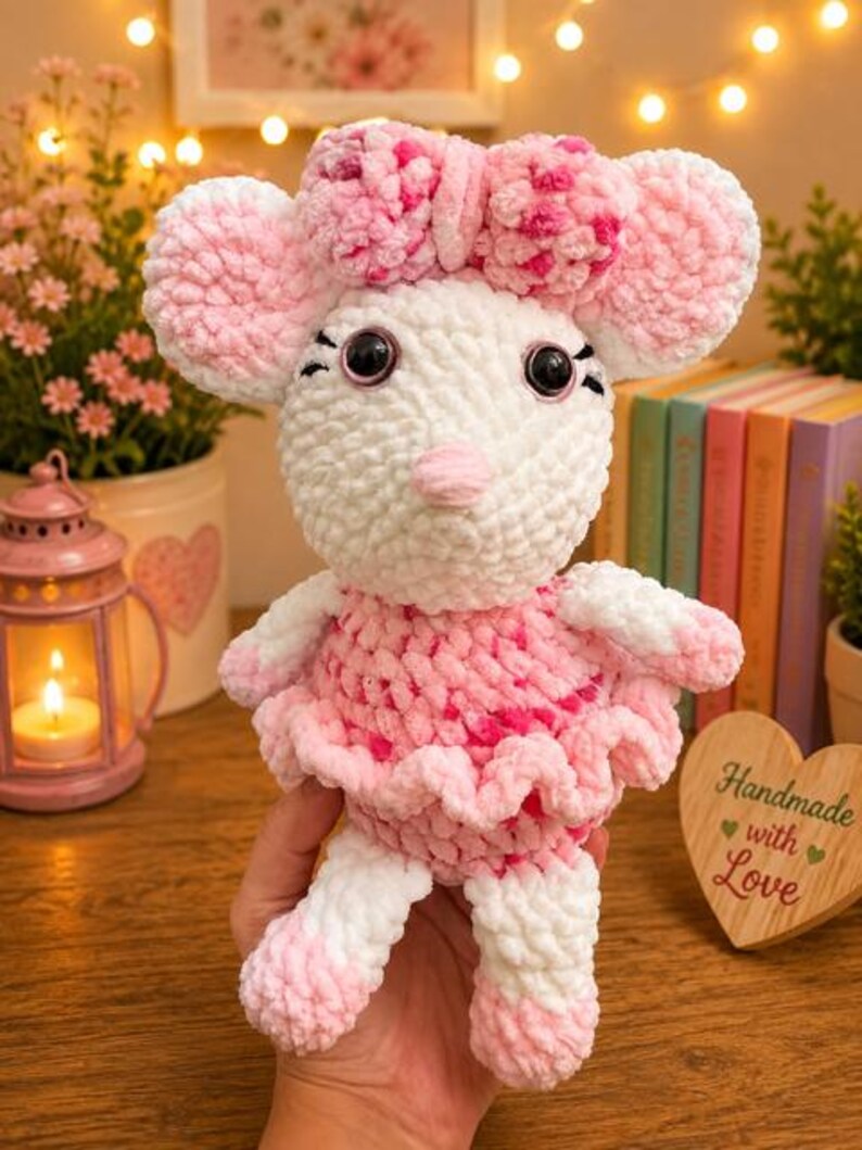 May include: A plush crochet mouse toy made of soft white and pink yarn. The mouse features large round ears, a pink nose, black plastic eyes, and a pink and white patterned bow on its head. It wears a ruffled pink and white skirt and has matching pink and white feet.