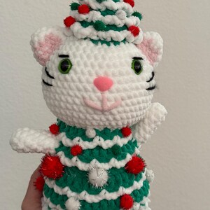 Pode incluir: Uma boneca de gato branca de croch&ecirc; usando um vestido e chap&eacute;u de &aacute;rvore de Natal verde e branco, adornada com pompons vermelhos e prateados. O gato tem orelhas internas rosa, nariz rosa, olhos verdes e bigodes pretos.