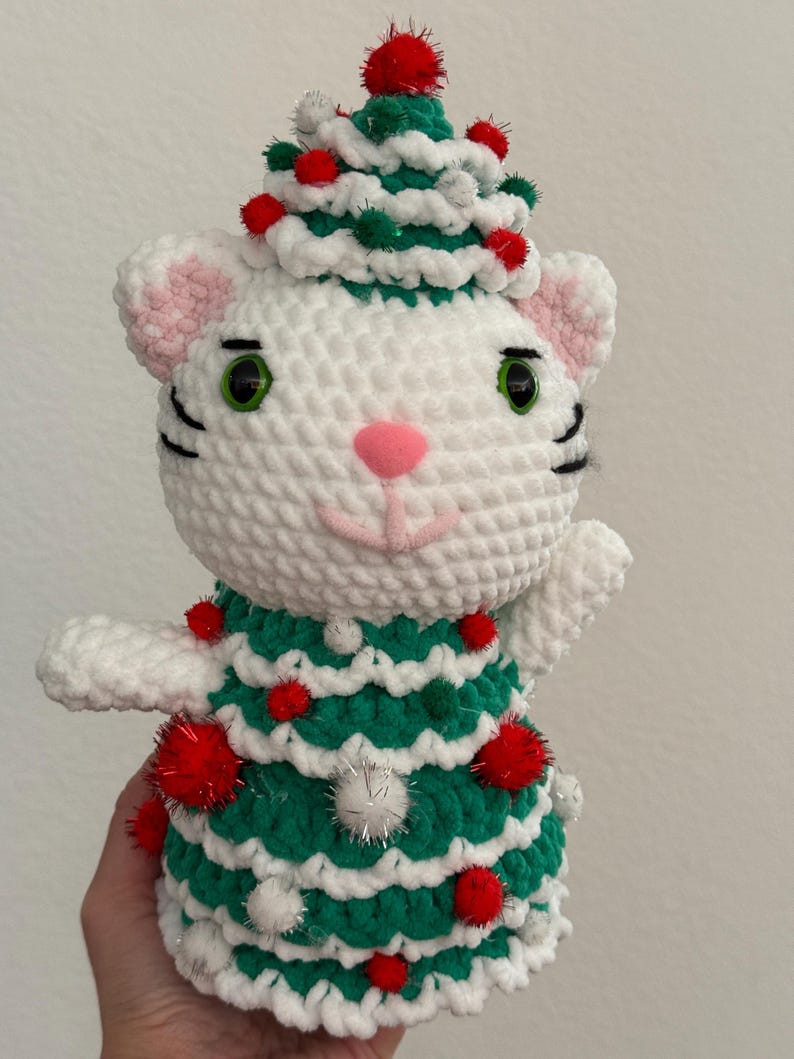 Pode incluir: Uma boneca de gato branca de croch&ecirc; usando um vestido e chap&eacute;u de &aacute;rvore de Natal verde e branco, adornada com pompons vermelhos e prateados. O gato tem orelhas internas rosa, nariz rosa, olhos verdes e bigodes pretos.
