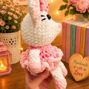 May include: A plush crochet toy shaped like a mouse with white and pink variegated yarn. The mouse features large ears, a ruffled skirt, and black plastic eyes. The soft, textured yarn creates a fuzzy appearance throughout the entire figure.