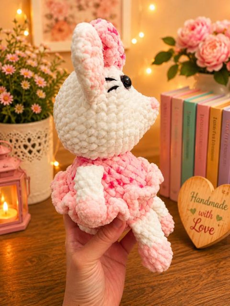 May include: A plush crochet toy shaped like a mouse with white and pink variegated yarn. The mouse features large ears, a ruffled skirt, and black plastic eyes. The soft, textured yarn creates a fuzzy appearance throughout the entire figure.