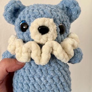 May include: A blue crocheted teddy bear with white crocheted trim around the neck. The bear has black eyes with blue glitter and a black nose.