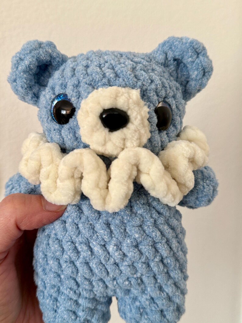 May include: A blue crocheted teddy bear with white crocheted trim around the neck. The bear has black eyes with blue glitter and a black nose.