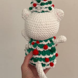 Pode incluir: Uma figura de gato branco de croch&ecirc; usando um vestido e chap&eacute;u de &aacute;rvore de Natal verde e vermelho. O chap&eacute;u &eacute; decorado com pompons vermelhos e verdes. O gato tem uma cauda branca e orelhas brancas pequenas. Feito &agrave; m&atilde;o.