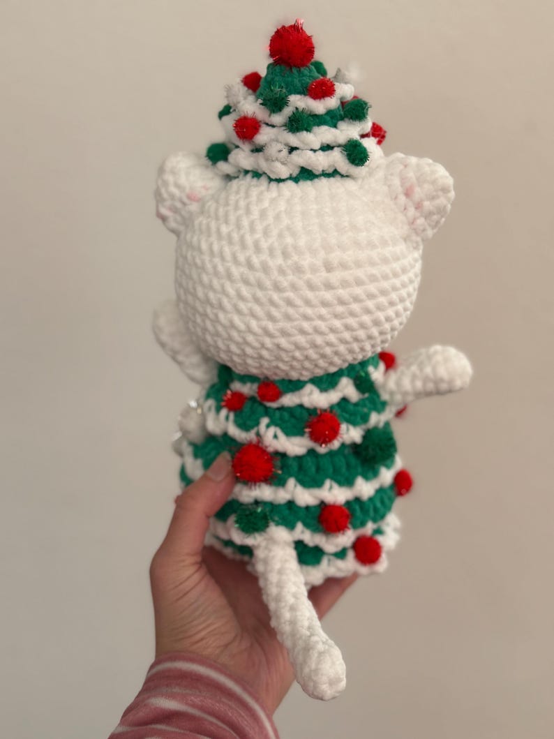 Pode incluir: Uma figura de gato branco de croch&ecirc; usando um vestido e chap&eacute;u de &aacute;rvore de Natal verde e vermelho. O chap&eacute;u &eacute; decorado com pompons vermelhos e verdes. O gato tem uma cauda branca e orelhas brancas pequenas. Feito &agrave; m&atilde;o.