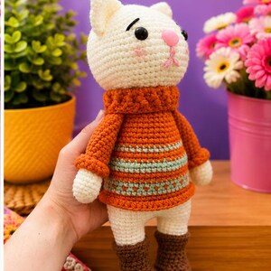 Pu&ograve; includere: Bambola a forma di gatto all'uncinetto fatta a mano con testa color crema, naso rosa e occhi neri. Indossa un maglione arancione con strisce blu, pantaloncini bianchi e stivali marroni. La bambola &egrave; tenuta in mano.