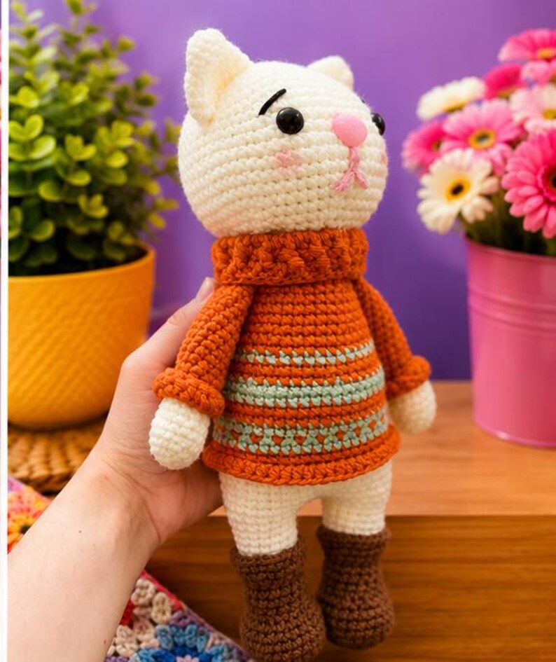 Pu&ograve; includere: Bambola a forma di gatto all'uncinetto fatta a mano con testa color crema, naso rosa e occhi neri. Indossa un maglione arancione con strisce blu, pantaloncini bianchi e stivali marroni. La bambola &egrave; tenuta in mano.