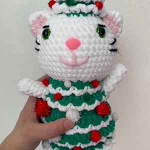Pode incluir: Boneca de gato de croch&ecirc; branco feita &agrave; m&atilde;o, vestindo um vestido e chap&eacute;u de &aacute;rvore de Natal verdes, adornada com pompons vermelhos e prateados. O gato tem orelhas e nariz rosa e olhos verdes. Uma decora&ccedil;&atilde;o festiva.