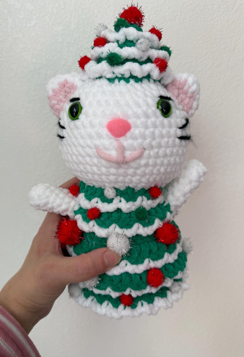Pode incluir: Boneca de gato de croch&ecirc; branco feita &agrave; m&atilde;o, vestindo um vestido e chap&eacute;u de &aacute;rvore de Natal verdes, adornada com pompons vermelhos e prateados. O gato tem orelhas e nariz rosa e olhos verdes. Uma decora&ccedil;&atilde;o festiva.
