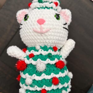 Pode incluir: Um gato de croch&ecirc; branco feito &agrave; m&atilde;o usando um vestido e chap&eacute;u de &aacute;rvore de Natal verdes, adornado com pompons vermelhos e brancos. O gato tem orelhas e nariz rosa, olhos verdes e bigodes pretos. Uma decora&ccedil;&atilde;o festiva de Natal.