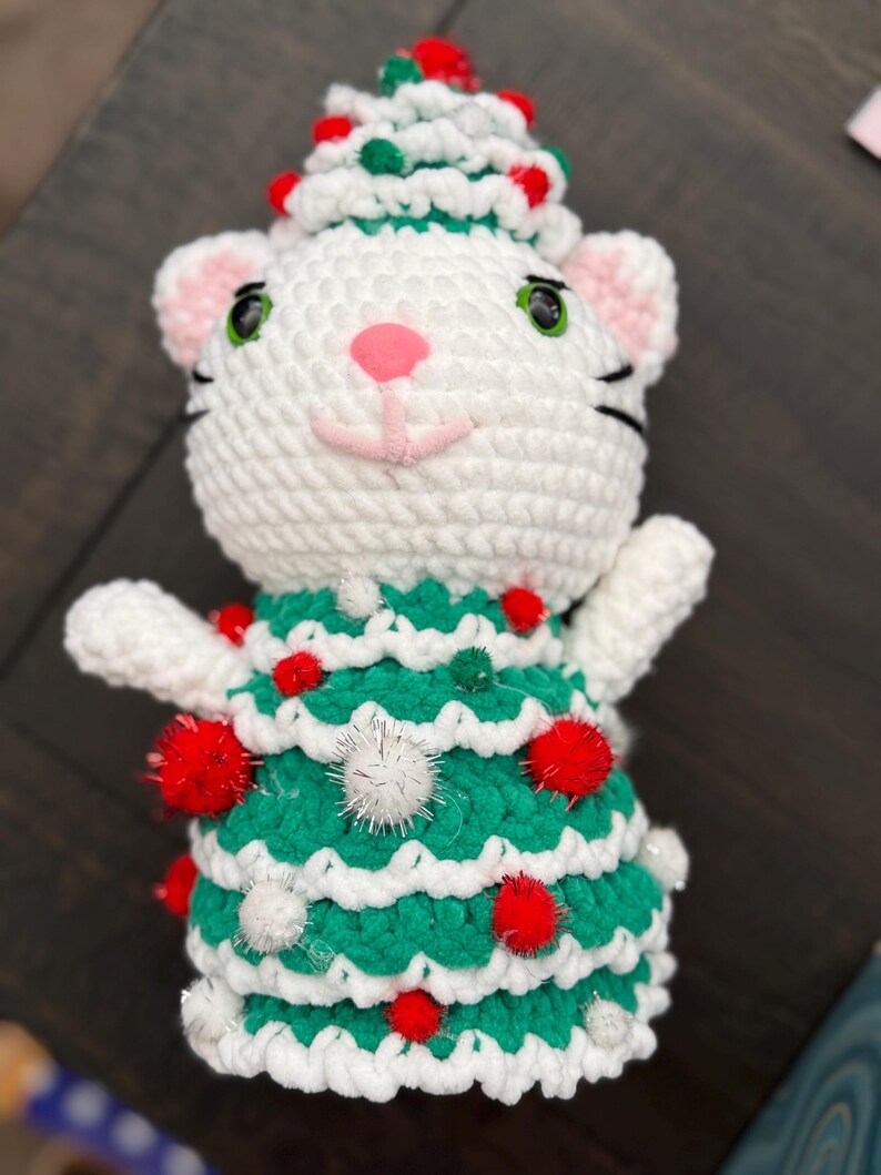 Pode incluir: Um gato de croch&ecirc; branco feito &agrave; m&atilde;o usando um vestido e chap&eacute;u de &aacute;rvore de Natal verdes, adornado com pompons vermelhos e brancos. O gato tem orelhas e nariz rosa, olhos verdes e bigodes pretos. Uma decora&ccedil;&atilde;o festiva de Natal.