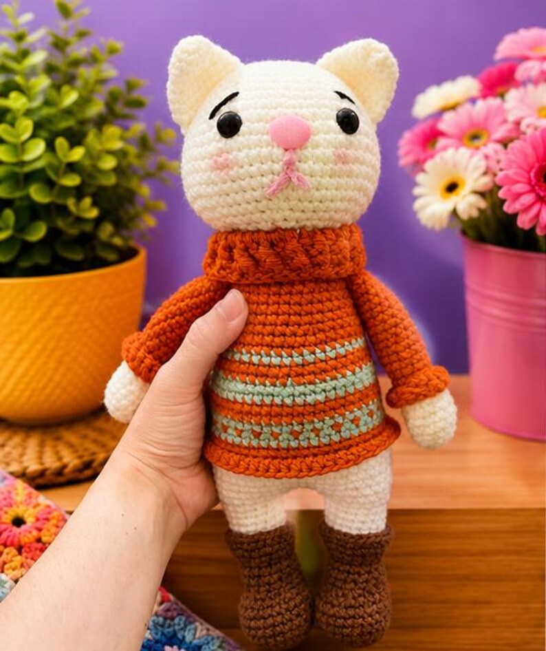 Pu&ograve; includere: Bambola a forma di gatto all'uncinetto fatta a mano con testa bianca, naso rosa e occhi neri. Il gatto indossa un maglione arancione con strisce blu, pantaloni bianchi e stivali marroni. La bambola &egrave; tenuta in mano, con fiori e una pianta sullo sfondo.