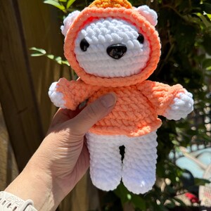 May include: A handmade white and orange crochet bear wearing a hoodie. The bear has black eyes and a black nose. The hoodie is orange with a lighter orange trim. The bear's paws and ears are white. The bear is a soft, plush toy.