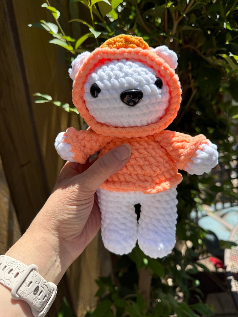 May include: A handmade white and orange crochet bear wearing a hoodie. The bear has black eyes and a black nose. The hoodie is orange with a lighter orange trim. The bear's paws and ears are white. The bear is a soft, plush toy.