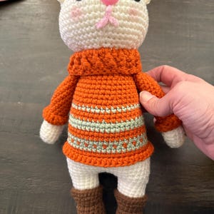 Pu&ograve; includere: Bambola a forma di gatto all'uncinetto fatta a mano con testa color crema, naso rosa e occhi neri. Indossa un maglione arancione con strisce verdi e bianche e stivali marroni. La bambola &egrave; alta circa 20 cm.