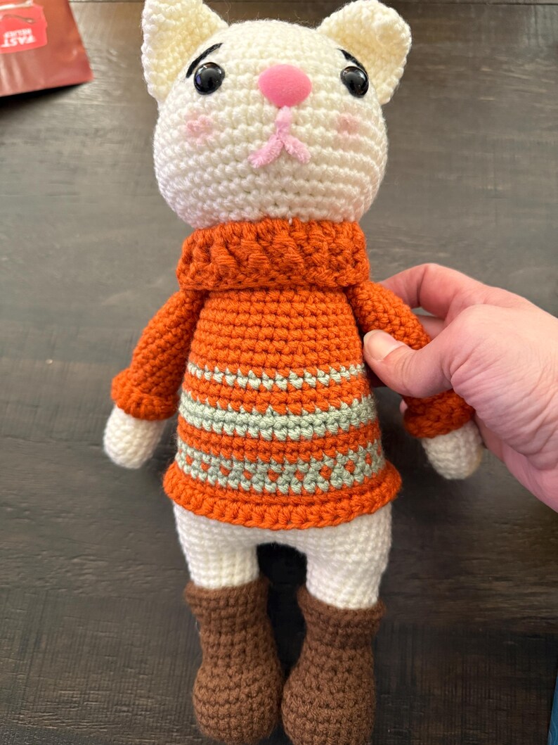 Pu&ograve; includere: Bambola a forma di gatto all'uncinetto fatta a mano con testa color crema, naso rosa e occhi neri. Indossa un maglione arancione con strisce verdi e bianche e stivali marroni. La bambola &egrave; alta circa 20 cm.