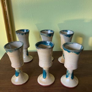 Puede incluir: Juego de seis copas de cerámica con base marrón claro y borde azul verdoso. Las copas tienen una copa redonda y un tallo alto y delgado.