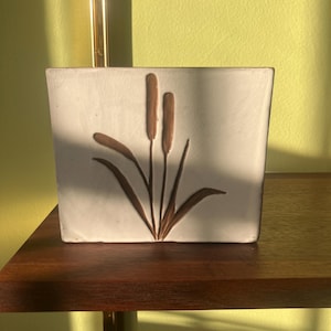 May include: A white ceramic tile with a brown raised design of three stylized reeds. The tile is sitting on a wooden shelf.