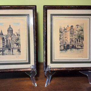May include: Two framed prints depicting Amsterdam canal scenes. Each print features a multi-layered matting with gold accents and a dark, mottled brown frame. The artwork shows historic buildings, bridges, and water, with handwritten text below.
