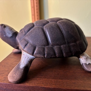 Może przedstawiać: Brązowa ceramiczna figurka żółwia z teksturowanym pancerzem i szczegółowymi nogami. Żółw stoi na drewnianej powierzchni, podkreślając swój rustykalny urok. Figurka ma szczegółowy wzór pancerza i wygląd postarzanego.