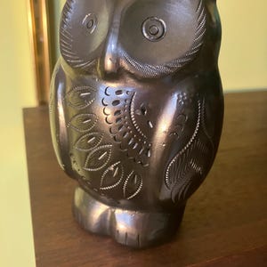 May include: A dark brown owl ornament with detailed carvings. The owl has large, round eyes and intricate feather patterns. The ornament is made of a smooth material and sits on a wooden shelf. The owl is a decorative item, possibly ceramic.