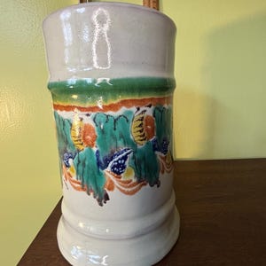 May include: A tall, off-white ceramic beer stein with a wide base and a cylindrical body. The stein features a band of green, orange, and yellow near the top, and a colorful floral design with blue and yellow accents. The stein is unglazed.