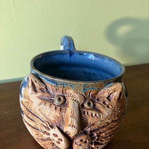 May include: A ceramic mug with a cat face sculpted on the front. The mug is glazed in shades of blue and brown. The cat's eyes are green and the nose is brown. The mug has a handle on the side.
