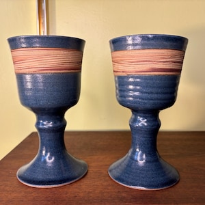 Puede incluir: Dos copas de cerámica azul con una banda de textura de madera marrón claro alrededor del centro. Las copas tienen una base de pedestal y un borde acampanado. El fondo es una pared amarillo claro y una superficie de madera marrón oscuro.