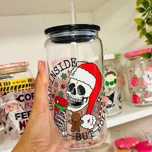 May include: A clear glass tumbler with a black lid and a straw. The tumbler has a festive design featuring a skull wearing a Santa hat and holding a cup of coffee. The text "Dead Inside But Jolly" is printed on the tumbler.
