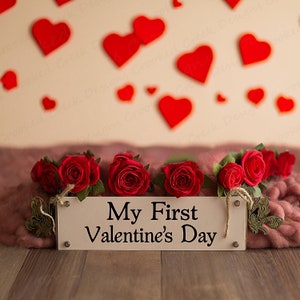 May include: A white wooden sign with the text "My First Valentine's Day" surrounded by red roses on a pink and brown background.