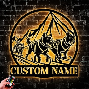 May include: A metal silhouette of two dogs pulling a sled with a mountain in the background. The silhouette is illuminated with a warm yellow light. The text "CUSTOM NAME" is below the silhouette.