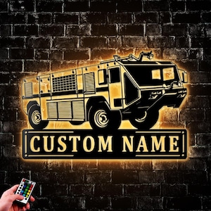 May include: A backlit sign featuring a detailed silhouette of a fire truck in black and gold. The sign includes the text "CUSTOM NAME" on a rectangular plaque. The background is a dark brick wall, and a remote control is visible.