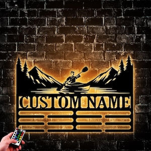 May include: A black metal wall decor with a silhouette of a person kayaking in a lake with mountains and trees in the background. The decor has a customizable nameplate that reads "CUSTOM NAME" and a row of slots for displaying items. The decor is illuminated with warm white LED lights.