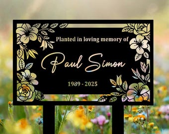 May include: Black memorial garden stake with floral cutouts and the text "Planted in loving memory of Paul Simon 1989-2025". The stake is set in a colorful flower garden.