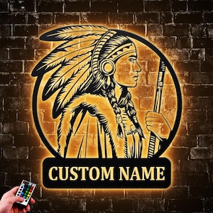 May include: A black and gold illuminated wall art piece featuring a Native American profile with a feathered headdress. The art is set within a circular frame, with the text "CUSTOM NAME" below. A remote control is visible in the lower left corner.