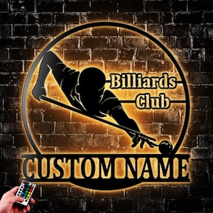 May include: A black metal billiards sign with a silhouette of a person playing pool, set within a circular frame. The sign includes the words "Billiards Club" and "CUSTOM NAME" with a remote control in the lower left corner.