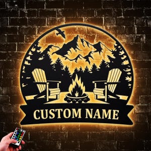May include: Black metal wall art with a mountain and forest scene, two Adirondack chairs, and a campfire. The words "CUSTOM NAME" are displayed on a banner. The art is illuminated with warm yellow light, and a remote control is visible.
