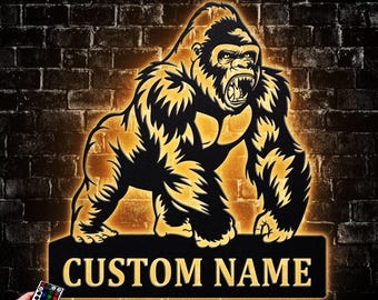 May include: A metal gorilla silhouette with a custom name plate. The gorilla is black with gold highlights and an open mouth. The sign is illuminated with a warm glow against a brick wall. A remote control is in the lower left corner.