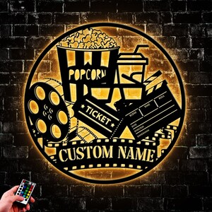 May include: A black metal movie night sign with a film reel, popcorn bucket, ticket, and clapperboard. The sign is illuminated with warm yellow light and has the text "CUSTOM NAME" on the bottom.