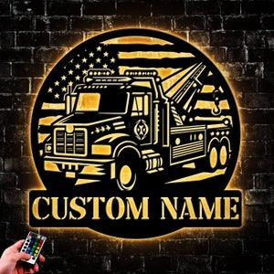 May include: A black metal silhouette of a tow truck with an American flag in the background. The tow truck has a hook and a flatbed. The silhouette is illuminated with a warm yellow light. The words "CUSTOM NAME" are below the silhouette.