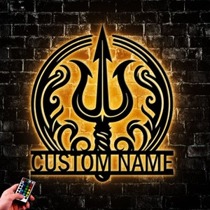 May include: A black metal wall decor with a circular design featuring a trident and floral accents. The design is illuminated with warm yellow light. The text "CUSTOM NAME" is displayed below the design.