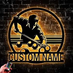 May include: A black metal silhouette of a person playing pool with a cue stick and billiard balls. The silhouette is set against a yellow brick wall background. The words "CUSTOM NAME" are displayed below the silhouette.
