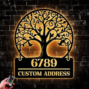 May include: A black metal tree of life design with a light shining through it. The tree is illuminated with warm white LED lights. The tree is attached to a black metal plaque with the numbers 6789 and the text "CUSTOM ADDRESS".