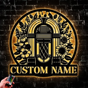 May include: A black and gold jukebox-themed wall sign with the words "CUSTOM NAME" at the bottom. The sign features a jukebox design with floral accents and musical notes. A remote control with multiple color options is held in the lower left corner.