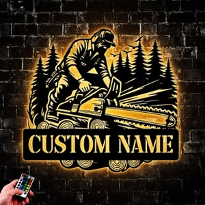 May include: A black and yellow metal silhouette of a person operating a chainsaw in a forest setting. The silhouette is illuminated with a warm yellow light. The text "CUSTOM NAME" is displayed on a black metal plaque below the silhouette.