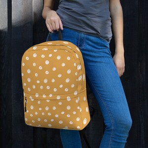May include: A mustard yellow backpack with a white polka dot pattern. The backpack has a black zipper and a black adjustable strap.