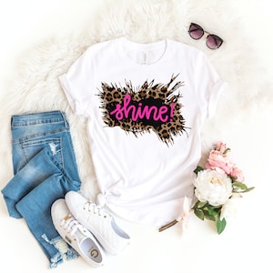 May include: White t-shirt with a black and brown leopard print design and the word "shine!" in pink lettering.