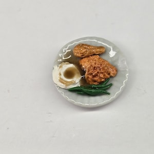 May include: A miniature plate featuring a meal of two pieces of golden-brown fried chicken, a scoop of white mashed potatoes topped with brown gravy, and a side of bright green beans, positioned next to a ruler showing imperial measurements.