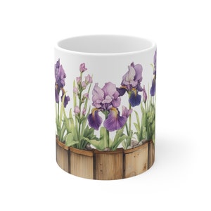 May include: White ceramic mug featuring a watercolor illustration of purple irises and other flowers in a wooden planter. The flowers have purple petals and green leaves. The mug has a smooth, glossy finish.