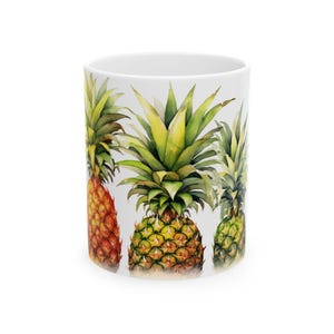 Pode incluir: Caneca de cerâmica branca com um desenho em aquarela de três abacaxis. Os abacaxis têm copas verdes e corpos amarelos e laranja texturizados. A caneca é cilíndrica e tem um acabamento brilhante.
